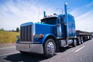 Classic blue bonneted big rig semi truck with chrome accessories and details on a multilane highway road with a laden flat bed trailer on background of blue sky.